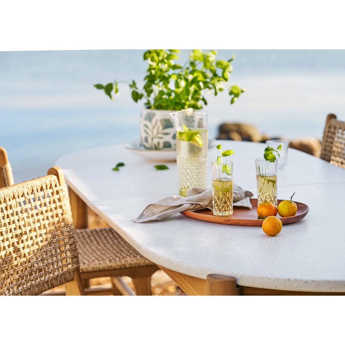 Outdoor dining table with glasses of lemonade, a tray of oranges, and a plant on a blurred beach background.