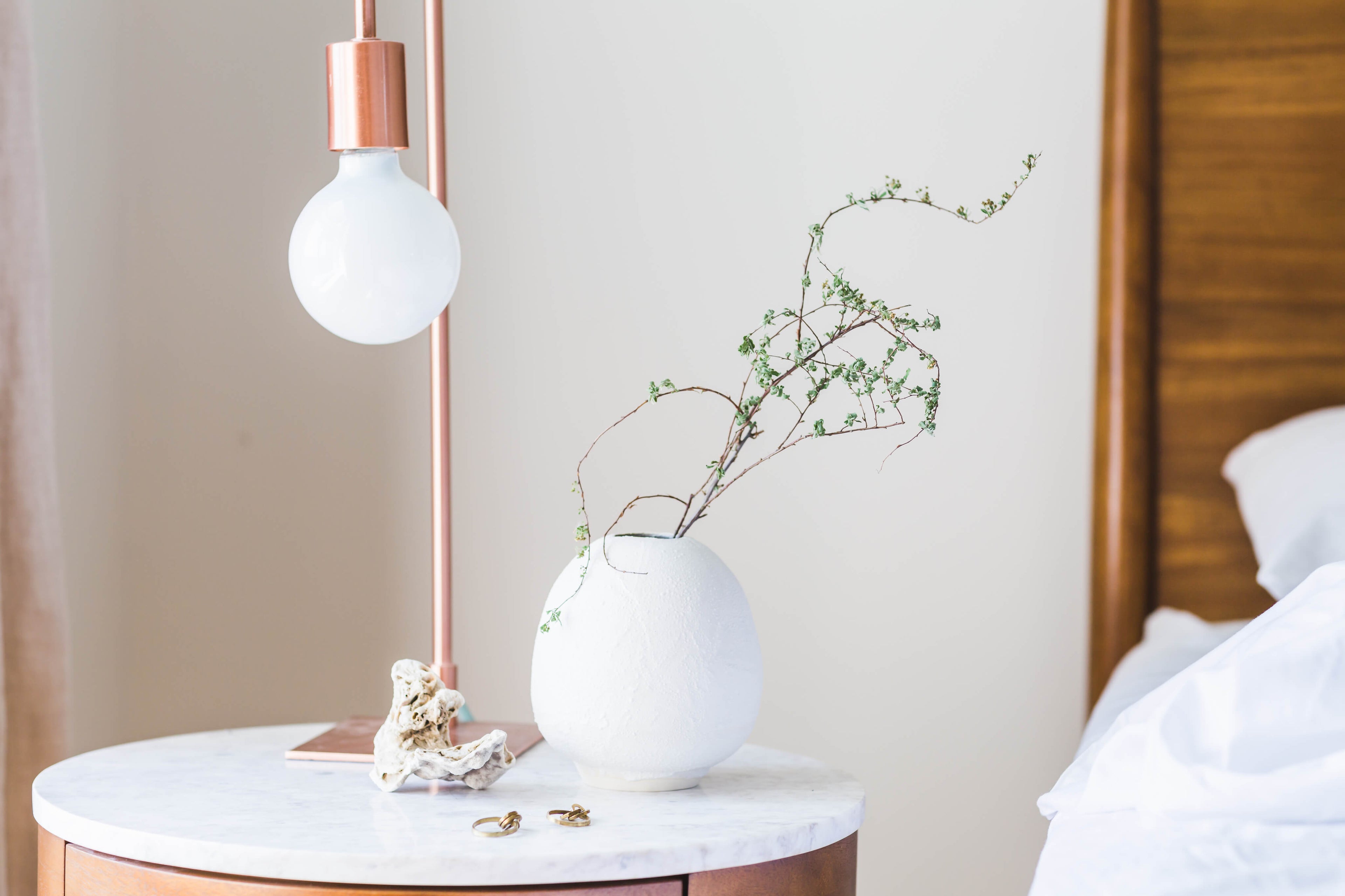 White vase with greenery on a marble side table next to a copper lamp.