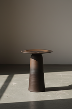 Tall ULFUR ceramic vase with wide conical base and flat disc top in bronze to black glaze on stone surface with soft daylight
