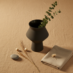 Overhead flat lay of the black speckled Cuzco ceramic vase on a sand linen surface with dried botanicals