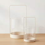 Set of two beige glass and metal hurricane candle holders on oak surface with soft daylight and clean off-white background