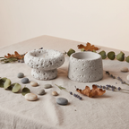Flat lay of two grey cement candle holders with linen cloth, pebbles and dried herbs on neutral background