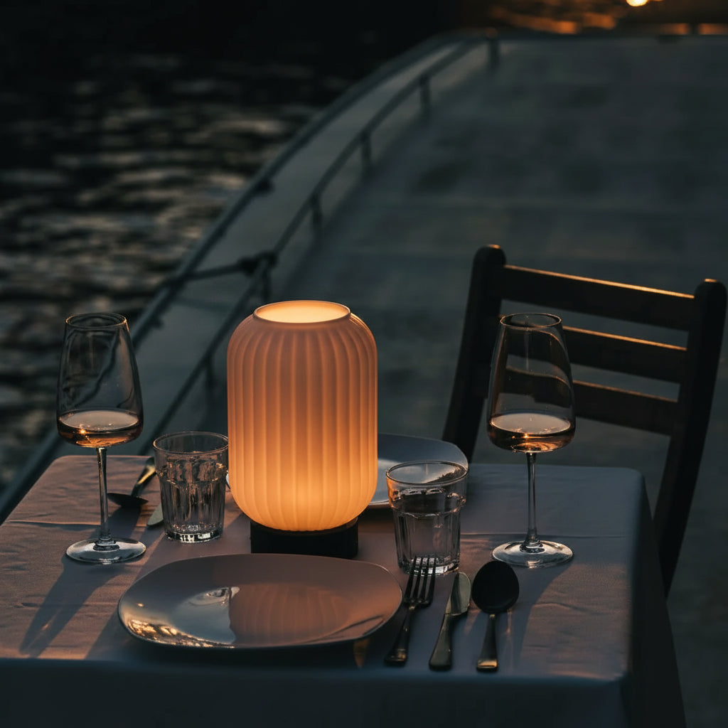 Dining table setting with wine glasses, lamp, and cutlery by a waterfront.